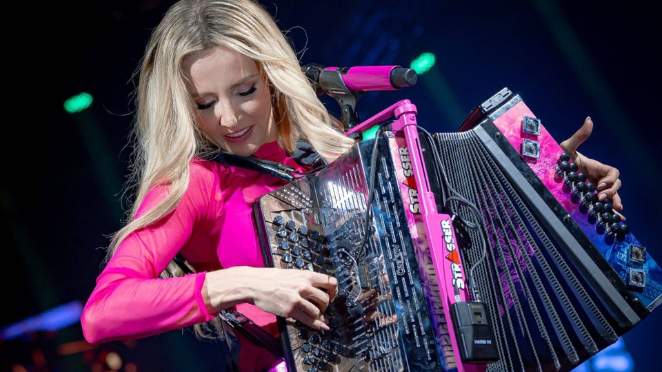 Melissa Naschenweng in an Konzert. Sie trägt pinkfarbene Kleidung und hält eine Harmonika in der Hand.