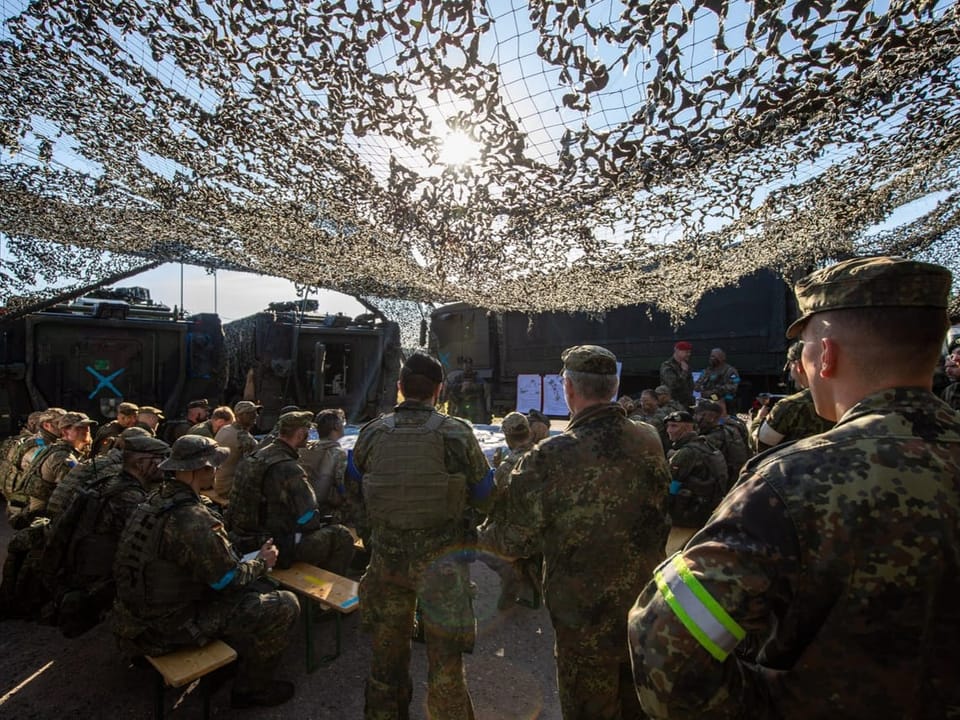 Soldaten in Tarnuniformen unter einem Tarnnetz im Freien.