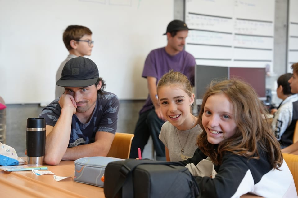 Mehrere Personen lachen und sprechen an einem Tisch in einem Klassenzimmer.