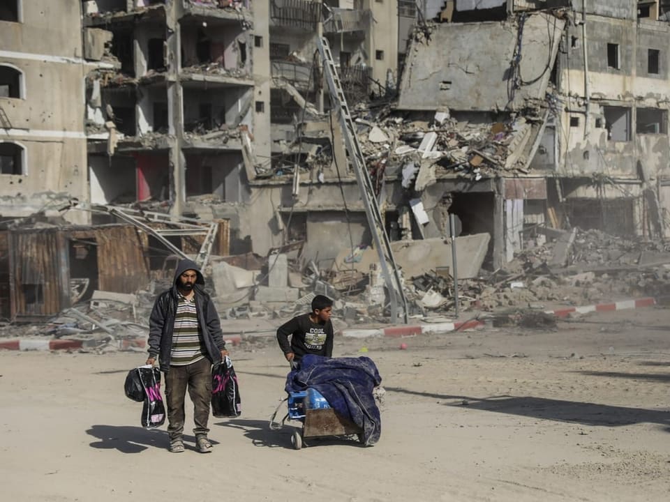 Zwei Palästinenser, ein Mann und ein Kind, laufen mit viel Gepäck an zerstörten Gebäuden vorbei.