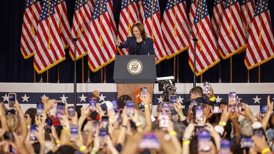 Person hält Rede auf der Bühne mit zahlreichen US-Flaggen, Publikum hebt Smartphones.
