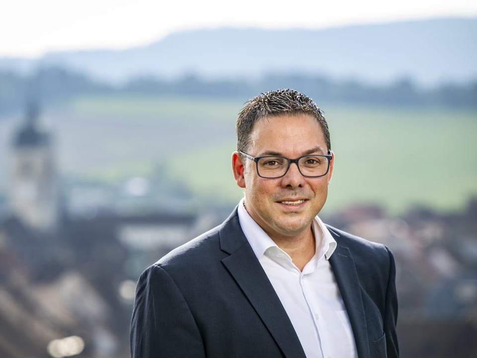 Mann mit Brille im Anzug vor unscharfem Landschaftshintergrund.