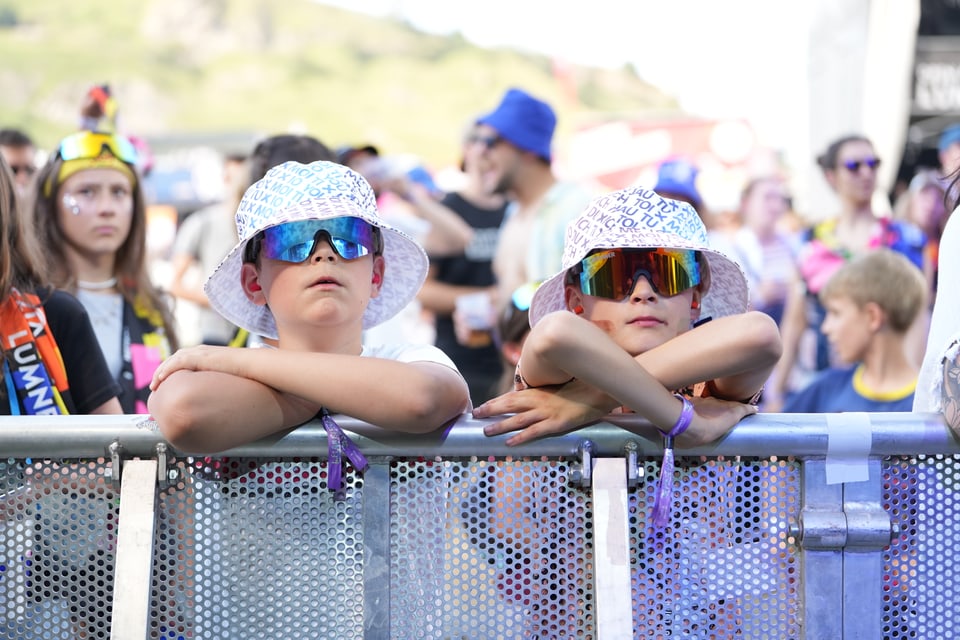 Kinder mit Sonnenbrillen und Hüten lehnen sich an ein Geländer bei einer Menschenmenge am Open Air Lumnezia 2024.