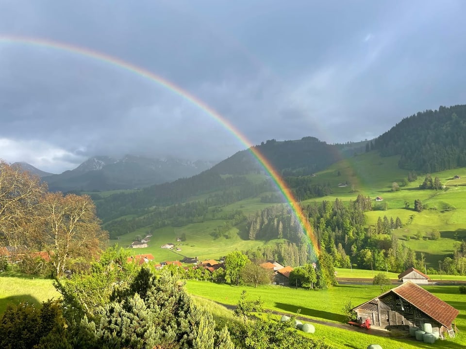 Regenbogen über grüner Landschaft mit Häusern und Bergen.