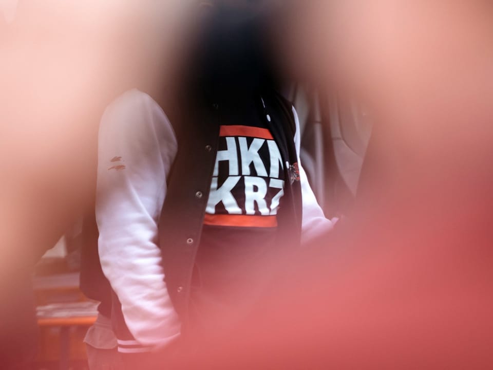 Vordergrund von Bild verschwommen, im Hintergrund Person mit T-shirt mit Aufdruck «HKNKRZ» zu erkennen.