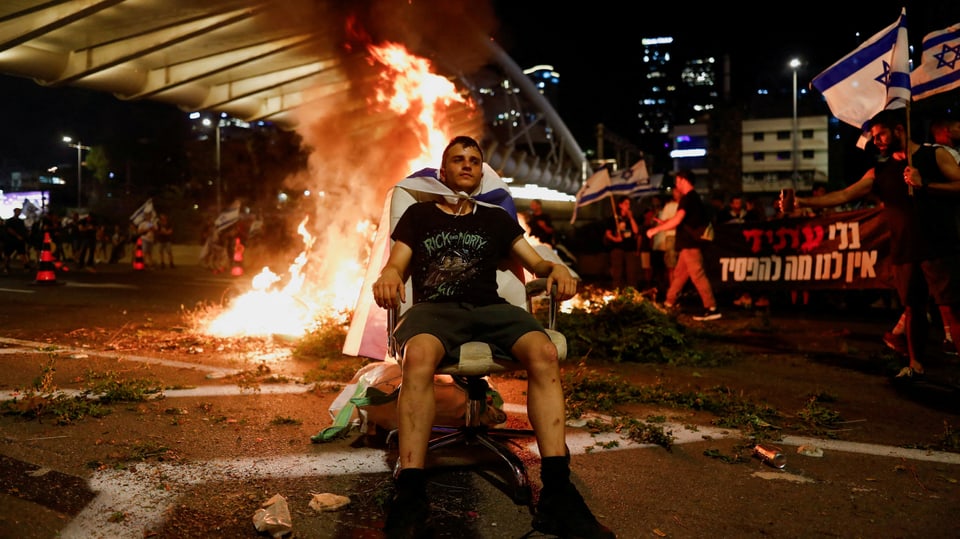 Ein Mann sitzt auf einem Stuhl auf einer Strasse vor einem Feuer. Er hat sich eine Israel-Fahne um den Hals gebunden.