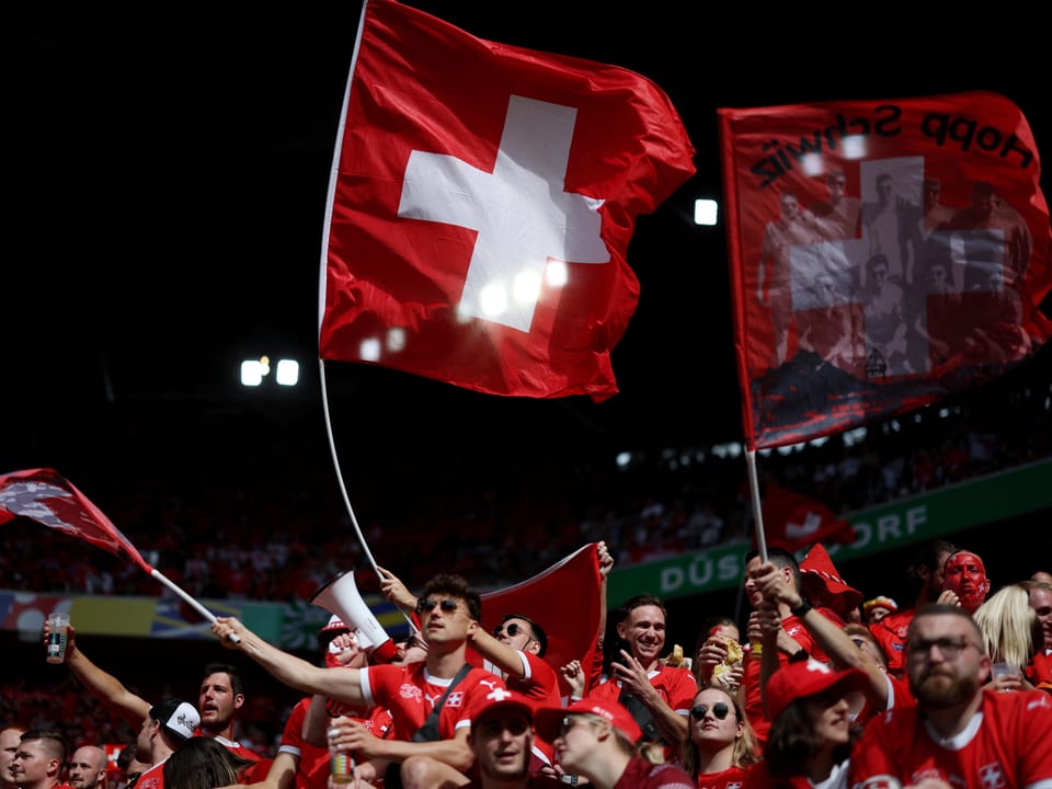 Eine Gruppe Menschen steht in Fussballtrikots und wehen Fahnen oder rufen etwas in ein Megafon. 