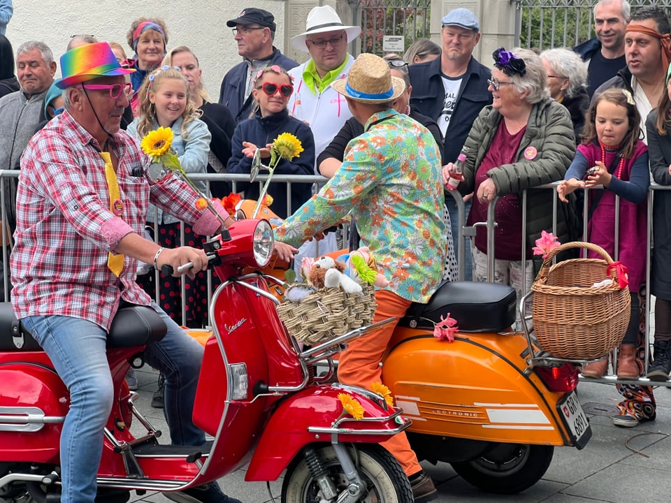 Zwei Männer in farbenfroher Kleidung auf Rollern werden von einer Menschenmenge beobachtet.