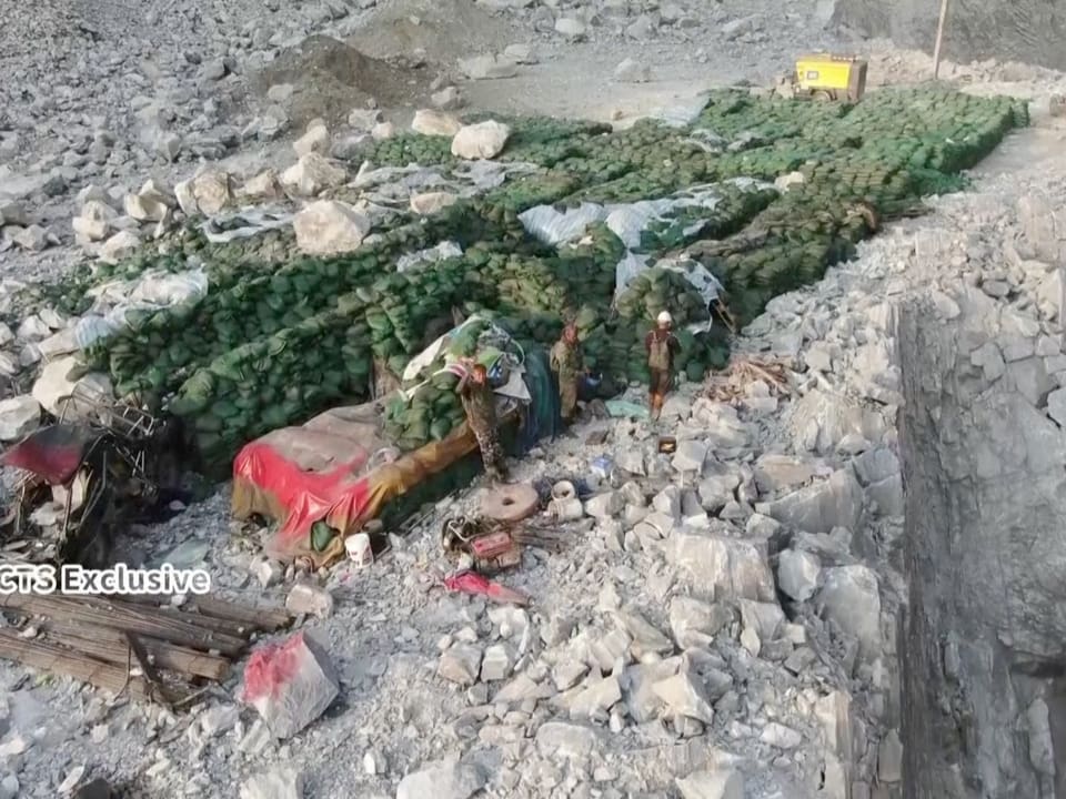 Foto einer Steinlandschaft, darauf viele grüne Säcke, drei Arbeiter zu sehen, stehen wartend an Säcke angelehnt.