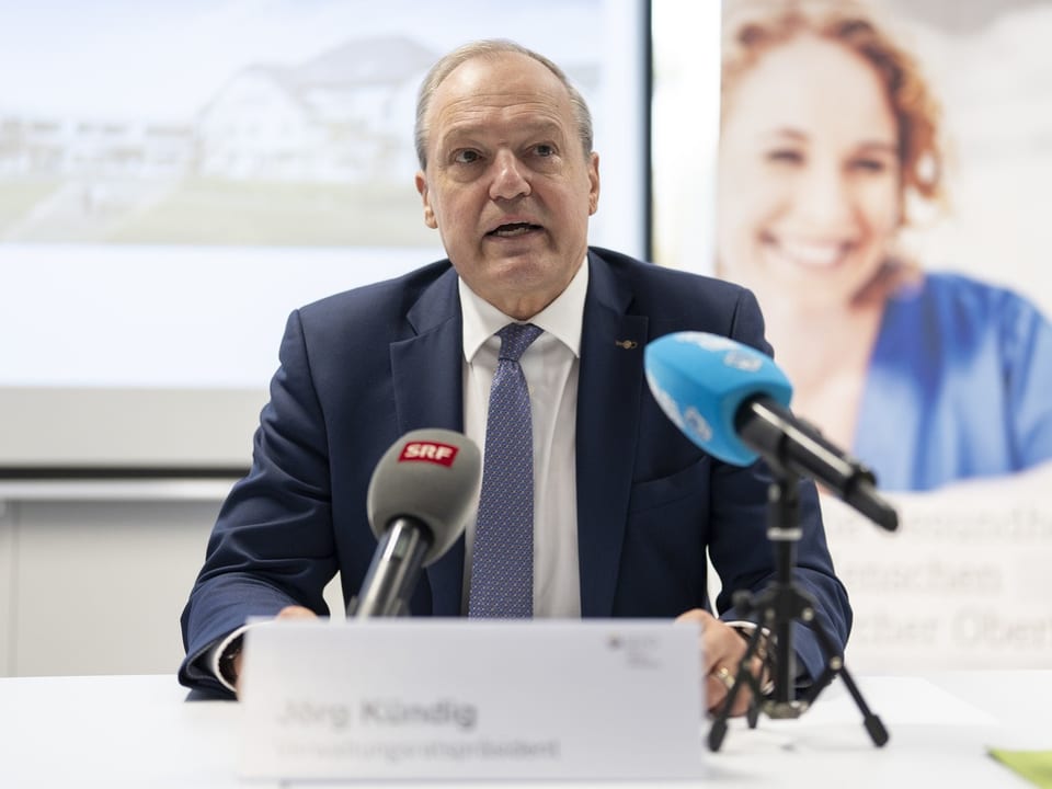 Mann im Anzug spricht bei Pressekonferenz mit Mikrofonen.