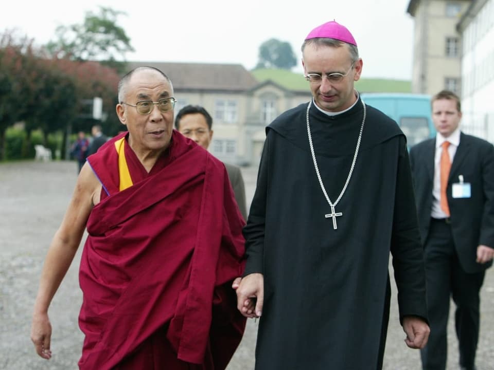 Ein buddhistischer Mönch und ein katholischer Geistlicher im Gespräch auf einem Innenhof.