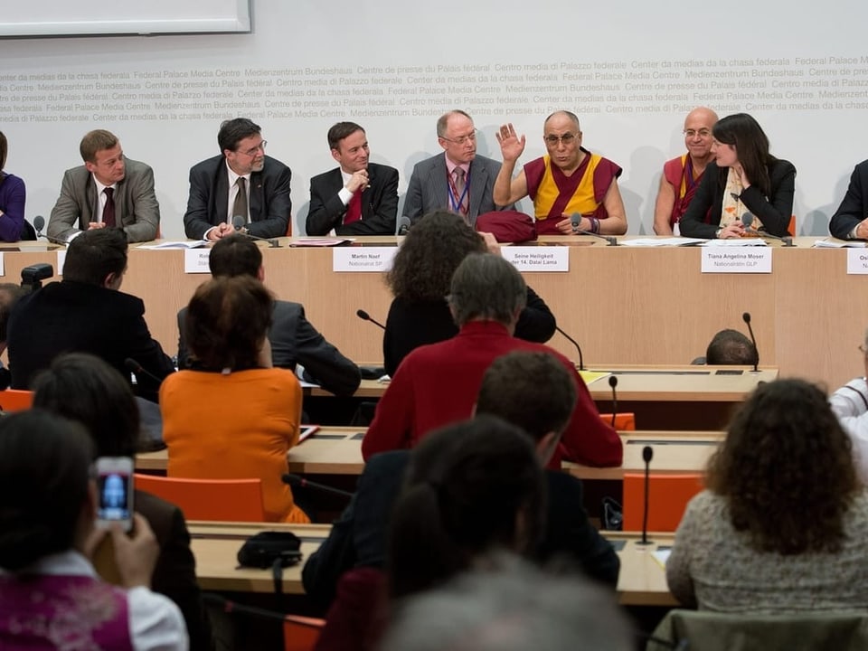 Menschen sitzen an einem Konferenztisch, ein Mann in gelboranger Robe spricht.