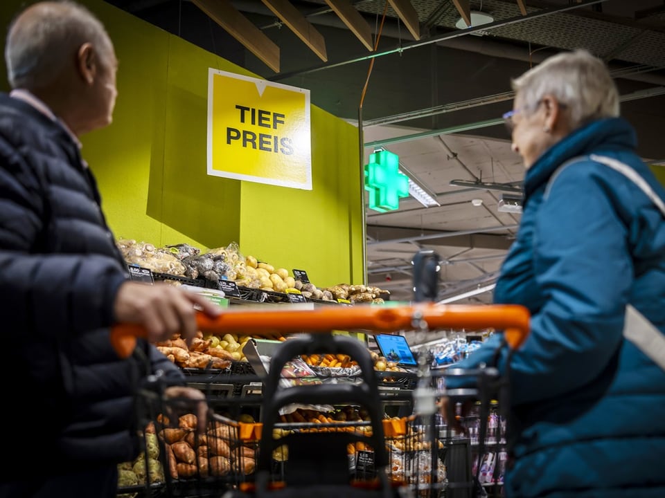 Zwei ältere Menschen stehen mit einem Einkaufswagen in der Migros. 