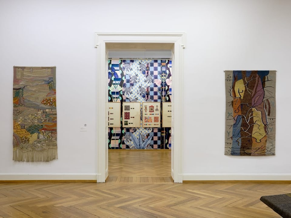 Galerieraum mit zwei Wandteppichen und Blick durch eine Tür auf ein buntes Wandmosaik.