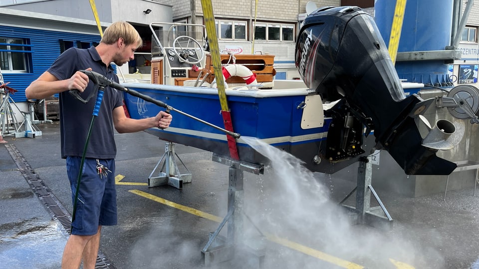 Ein Mann hälft einen Hochdruckreiniger. Aus diesem spritzt Wasser auf ein blaues Motorboot.