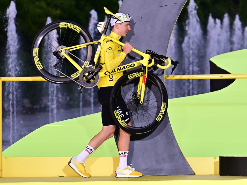 Radfahrer in gelber Kleidung trägt sein Fahrrad auf der Schulter vor einem Brunnen.