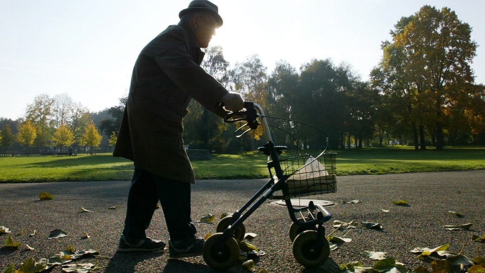 Ein älterer Herr mit Hut geht mit Hilfe eines Rollators durch einen Park.