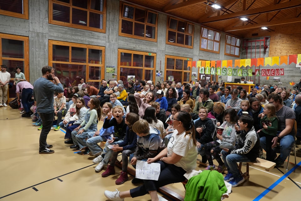 Volle Turnhalle mit Zuschauern auf Bänken, bunte Banner an der Wand.