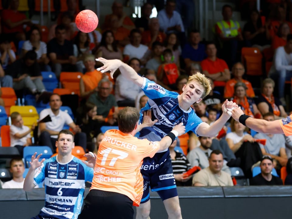 Handballspiel mit Spielern in blauen und orangefarbenen Trikots und einer jubelnden Menschenmenge im Hintergrund.