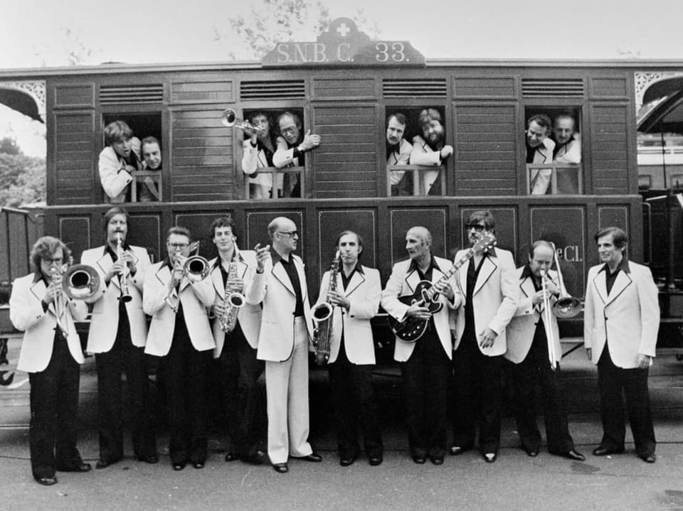 Bigband spielt vor historischem Eisenbahnwaggon, Musiker schauen aus Fenstern.