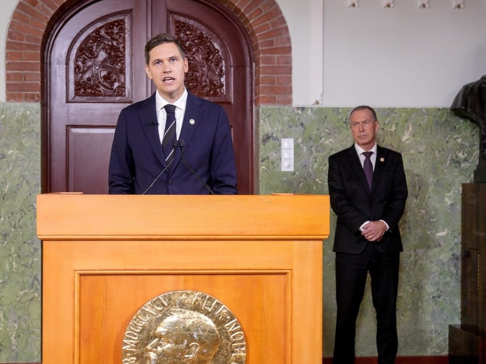 Mann hält Rede an Podium, zweiter Mann steht im Hintergrund.