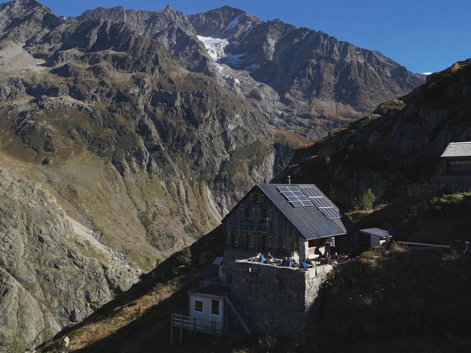 Drohnenaufnahme von einer Berghütte. Auf der Terrasse sitzen Menschen.