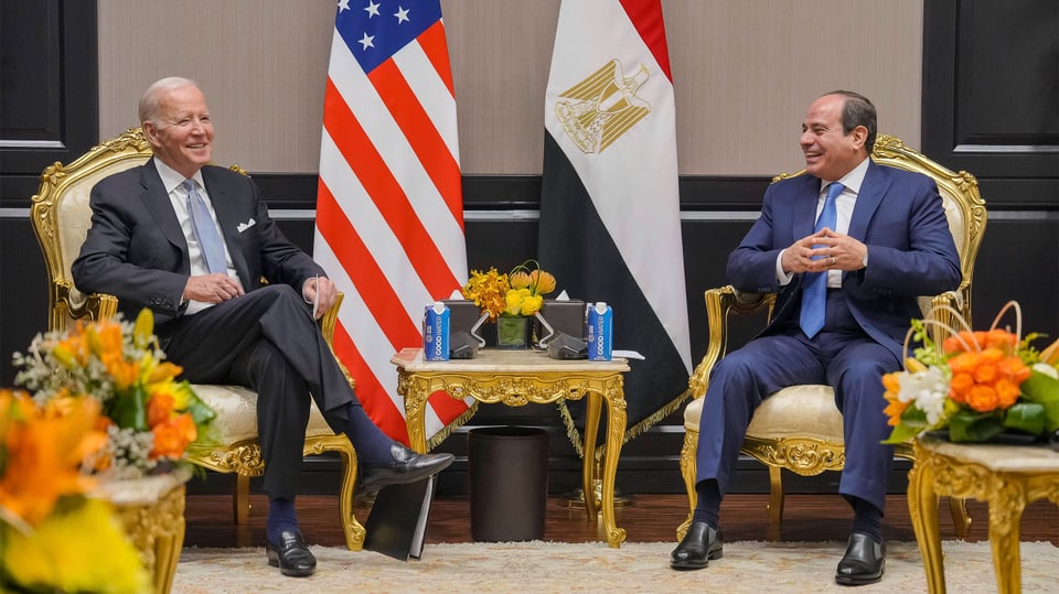 Zwei Männer in Anzügen sitzen auf goldenen Stühlen. Dazwischen und etwas dahinter stehen Flaggen.