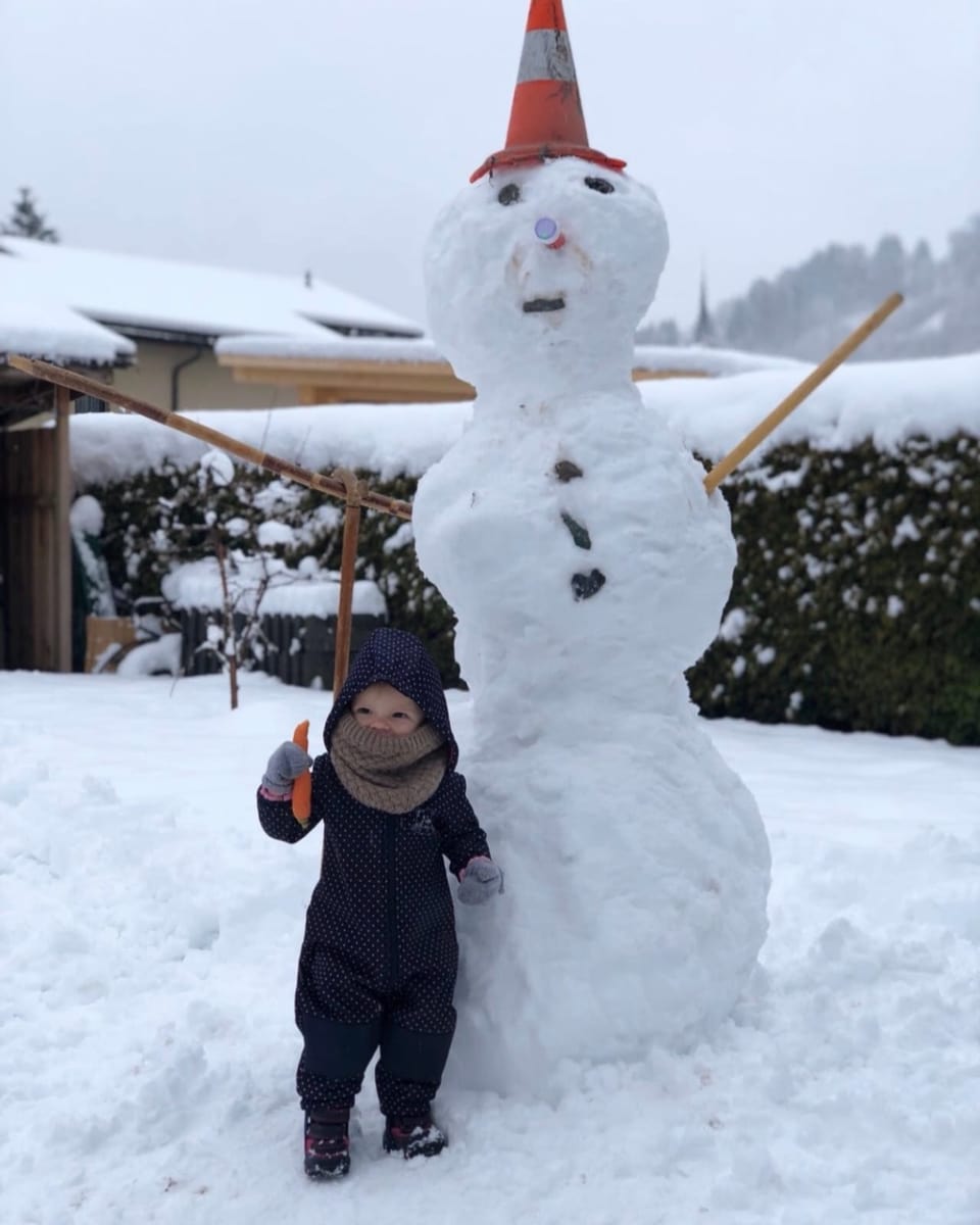 Schneemann mit Pilone auf dem Kopf. Daneben steht ein kleines Kind.