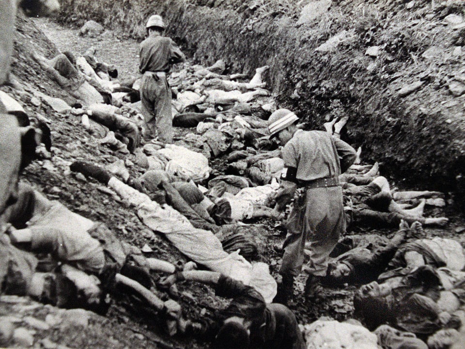 Soldaten gehen in einem Graben zwischen Leichen.