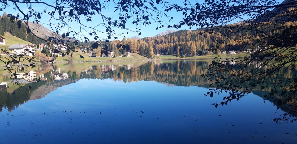 See mit Bergdorf und Herbstwald im Hintergrund.