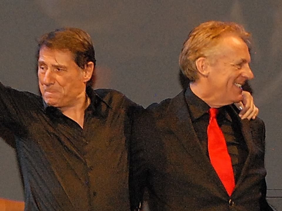 Udo Jürgens und Pepe Lienhard tragen beide schwarze Outfits und stehen auf der Bühne. Jürgens winkt, Lienhard lacht. 