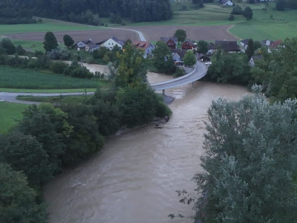 Luftaufnahme eines schlammigen Flusses mit Brücke und Dorf im Hintergrund.