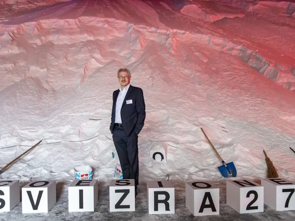 Mann steht vor Schneewand mit Buchstabenblöcken 'SVIZRA 27'