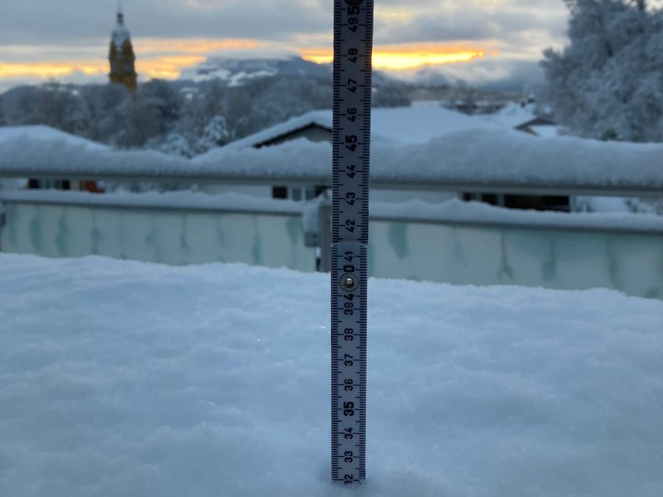 Lineal im Schnee misst Schneetiefe, Winterlandschaft im Hintergrund.