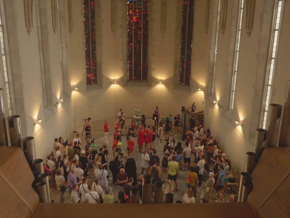 Der Kirchenraum ist gefüllt mit unterschiedlich bunt gekleideten Menschen.