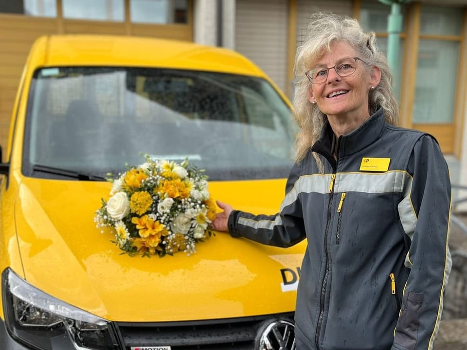Person in Arbeitskleidung steht neben gelbem Fahrzeug mit Blumenstrauss auf der Motorhaube.