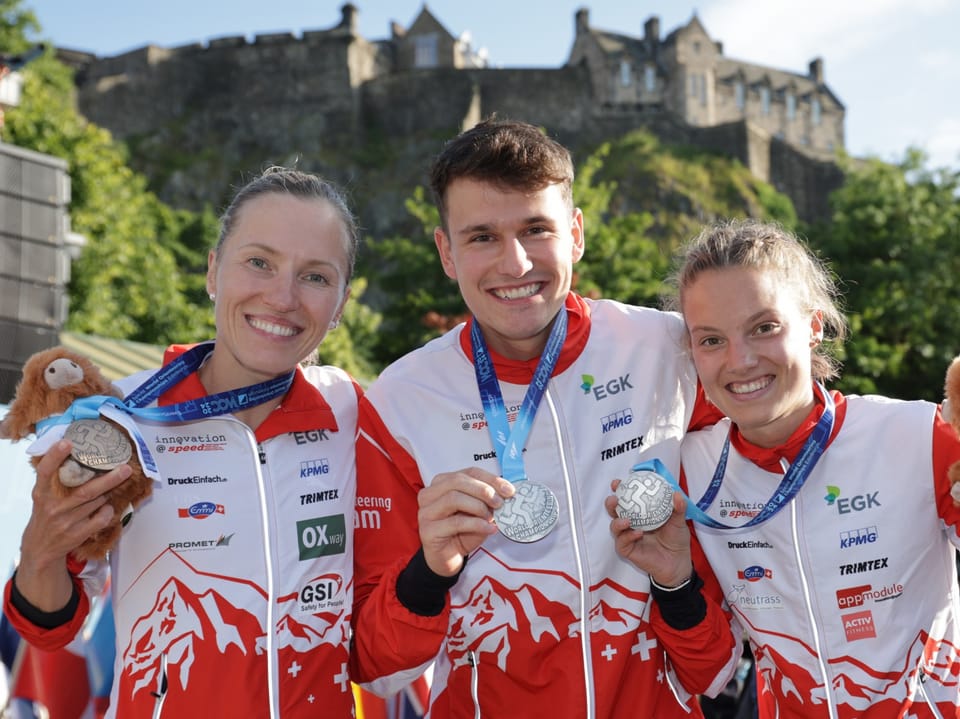 Drei lächelnde Athleten mit Medaillen vor einer Burg.