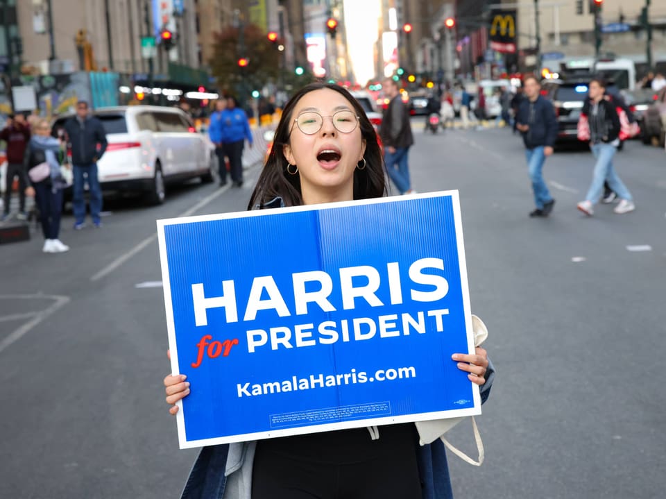 Person hält ein Schild mit 'Harris for President' auf einer geschäftigen Strasse.