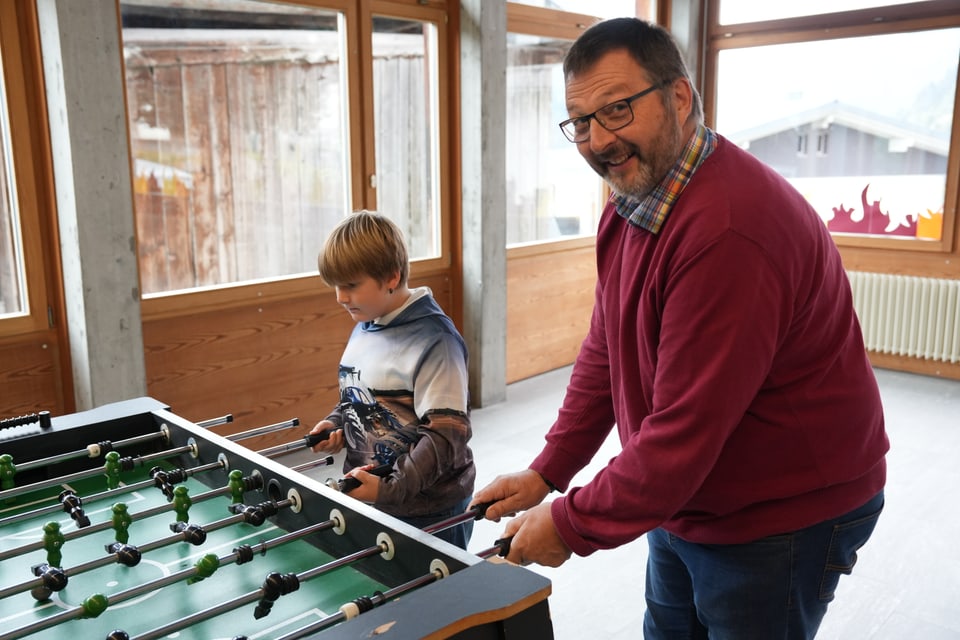 Mann und Junge spielen Tischfussball in einem Raum mit grossen Fenstern.