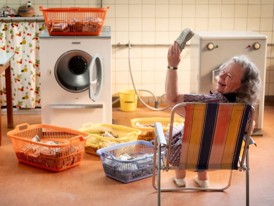 Ältere Frau sitzt lachend vor einer Waschmaschine mit Wäschekörben.