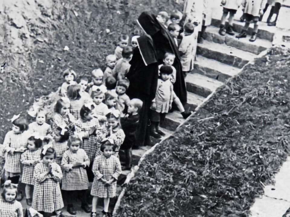 Schwarz-Weiss-Foto von Nonnen, die mit Kindern eine Treppe hinaufgehen.