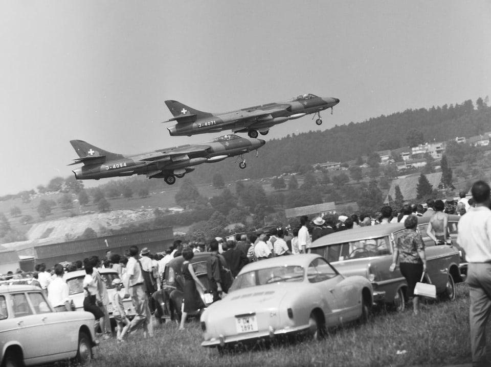 Zwei Kampfflugzeuge fliegen über eine Zuschauermenge, im Vordergrund sind parkierte Autos. 