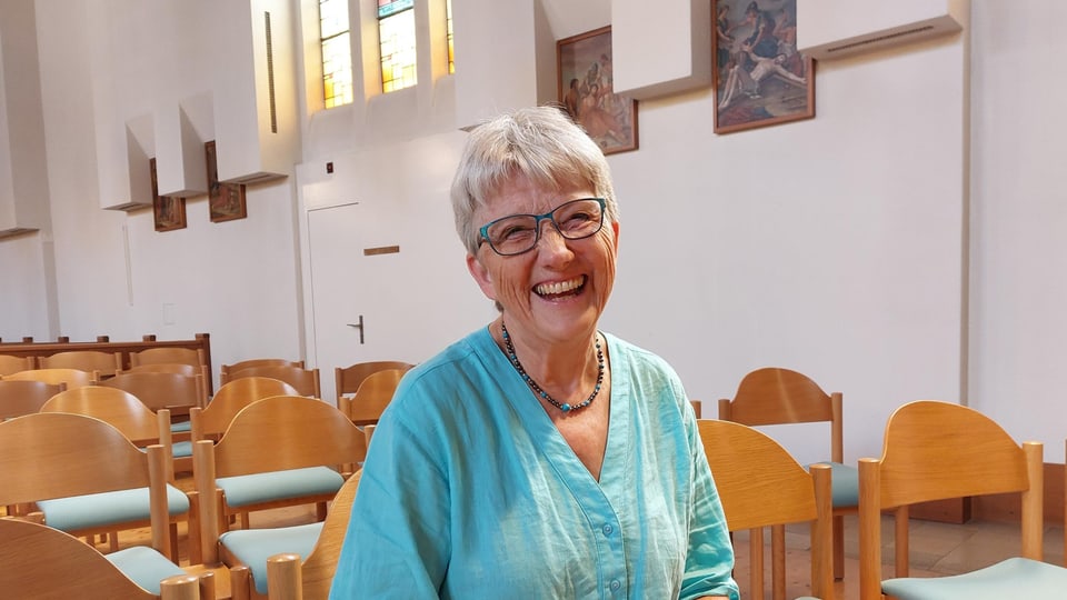 Eine alte Frau mit weissen, kurzen Haaren und Brille sitzt in einer Kirche und lacht.