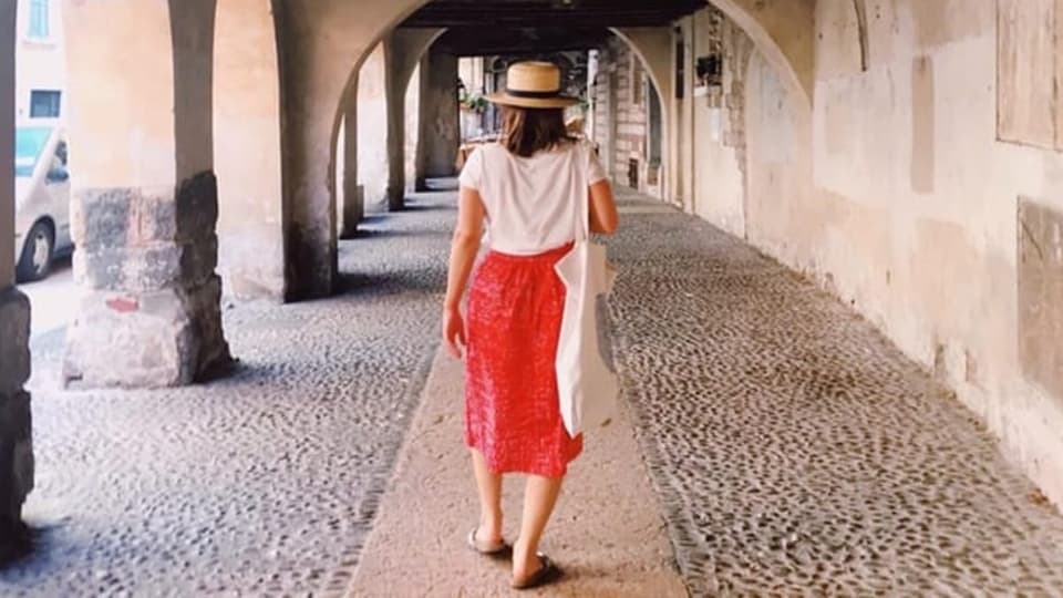 Eine Frau mittleren Alters mit Hut und rotem Jupe von hinten, wie sie durch eine Gewölbe-überdachte Strasse spaziert.