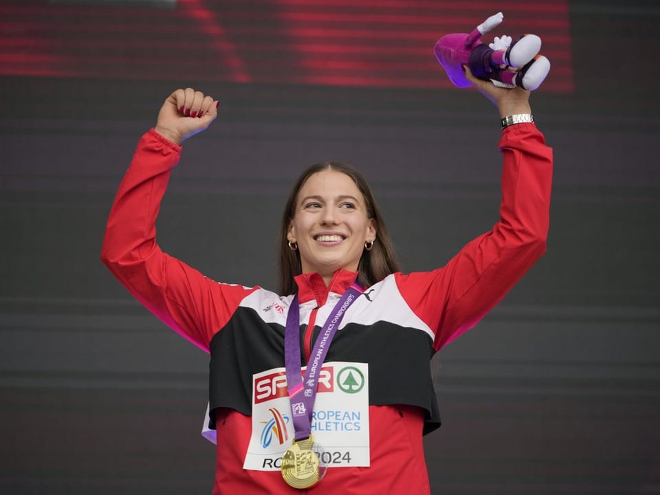 Frau im roten Anzug mit Medaille bei einer Siegerehrung, Arme erhoben.