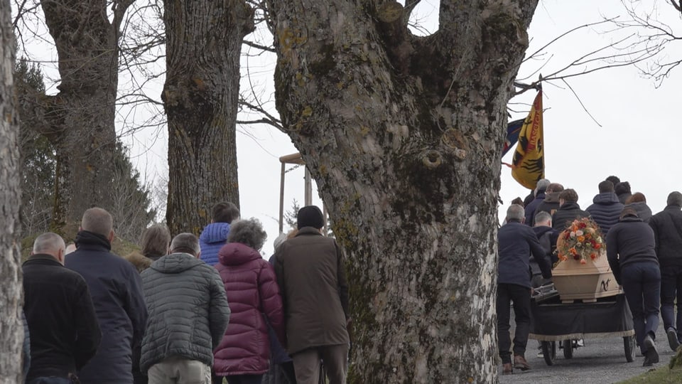 Trauergemeinde folgt Sarg auf Anhänger auf einem Weg unter Bäumen.