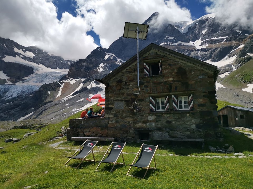 Steinhaus in den Bergen mit Sonnenliegen im Vordergrund und Personen unter Schirmen.