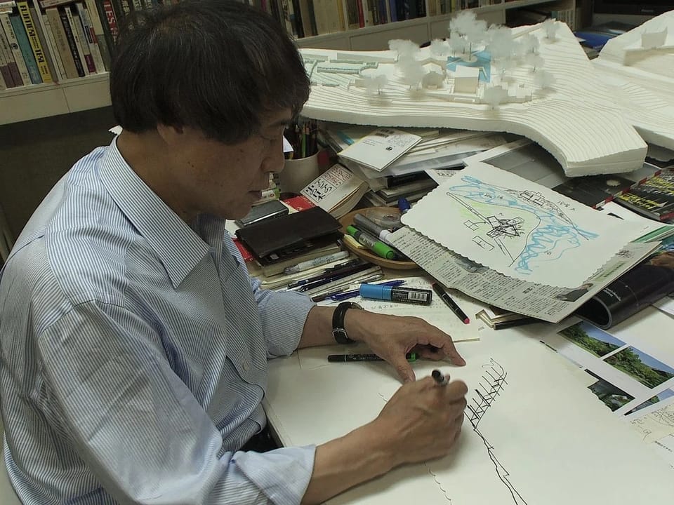 Mann zeichnet an einem Schreibtisch mit Architekturmodellen und Zeichnungen.