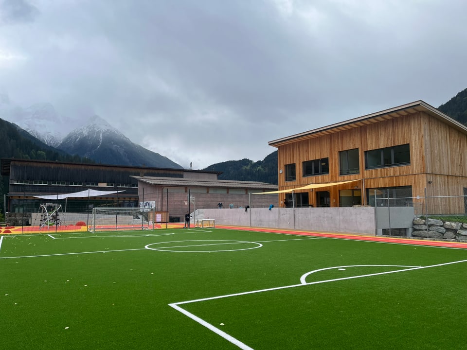 Fussballplatz mit Gebäuden und Bergen im Hintergrund.