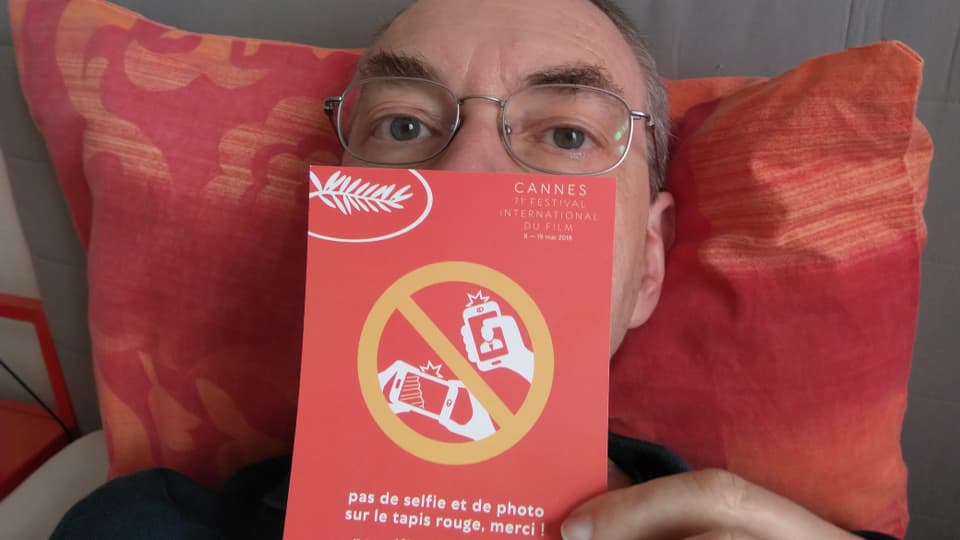Ein Mann liegt auf einem roten Kissen, er trägt eine Brille und hält einen Festival-Flyer von Cannes in der Hand.